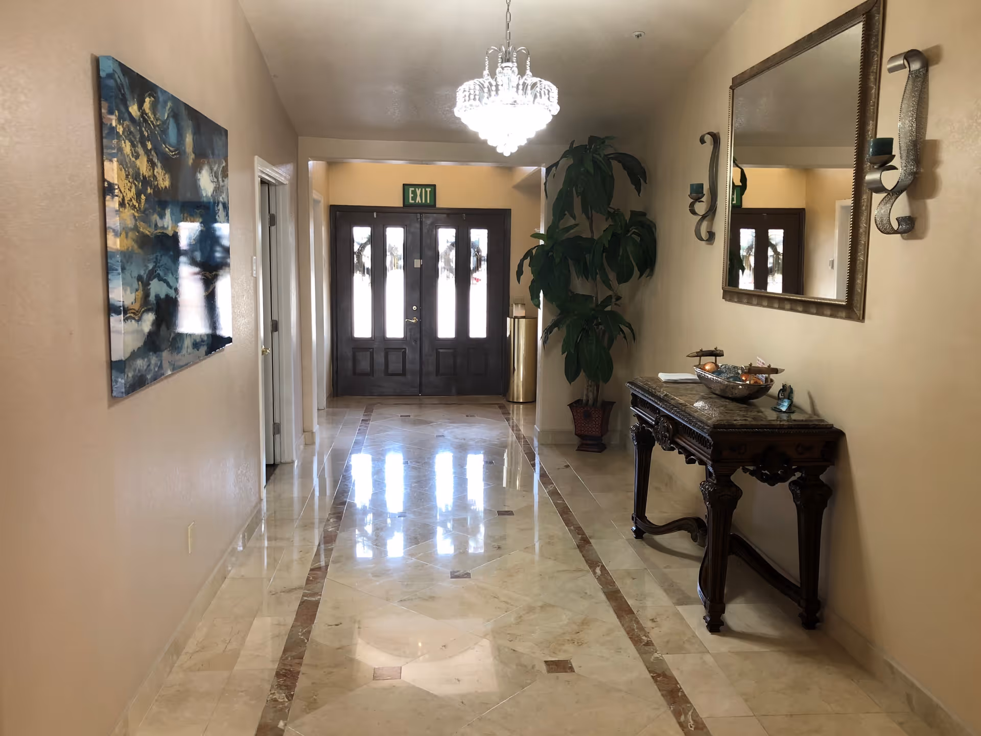 A polished marble floor hallway with beige walls, a decorative wooden table with a bowl and small items on the right, a large mirror and two wall-mounted candle holders above it, a large potted plant near the double glass doors with an exit sign above, and an abstract painting on the left wall. A chandelier hangs from the ceiling.