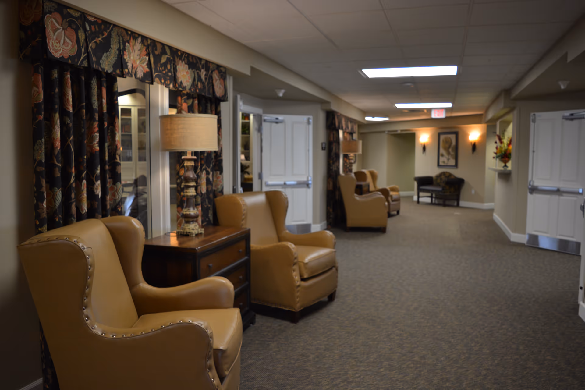 A hallway in Edgemont Place featuring several tan leather armchairs with nailhead trim, dark wooden side tables with lamps, floral patterned curtains, and soft lighting from ceiling fixtures and wall sconces. The carpeted floor and neutral wall colors create a warm and inviting atmosphere.