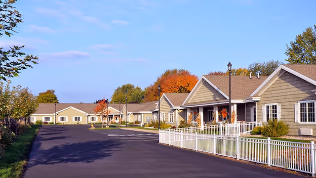 Exterior view of a senior living facility with single-story buildings featuring beige siding and white trim, a paved driveway, white fencing, and trees with autumn foliage under a clear blue sky.