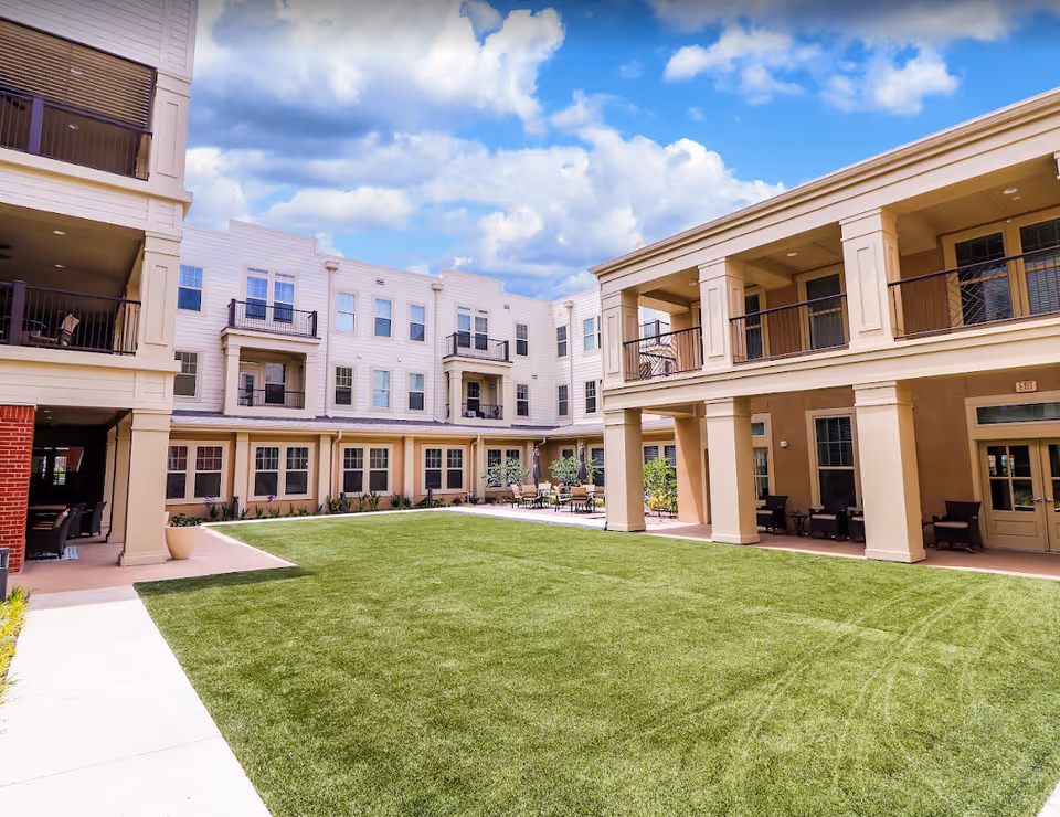 Outdoor courtyard area of Brigham House senior living facility with well-maintained green lawn, surrounded by multi-story buildings featuring balconies and seating areas under covered walkways, under a partly cloudy blue sky.