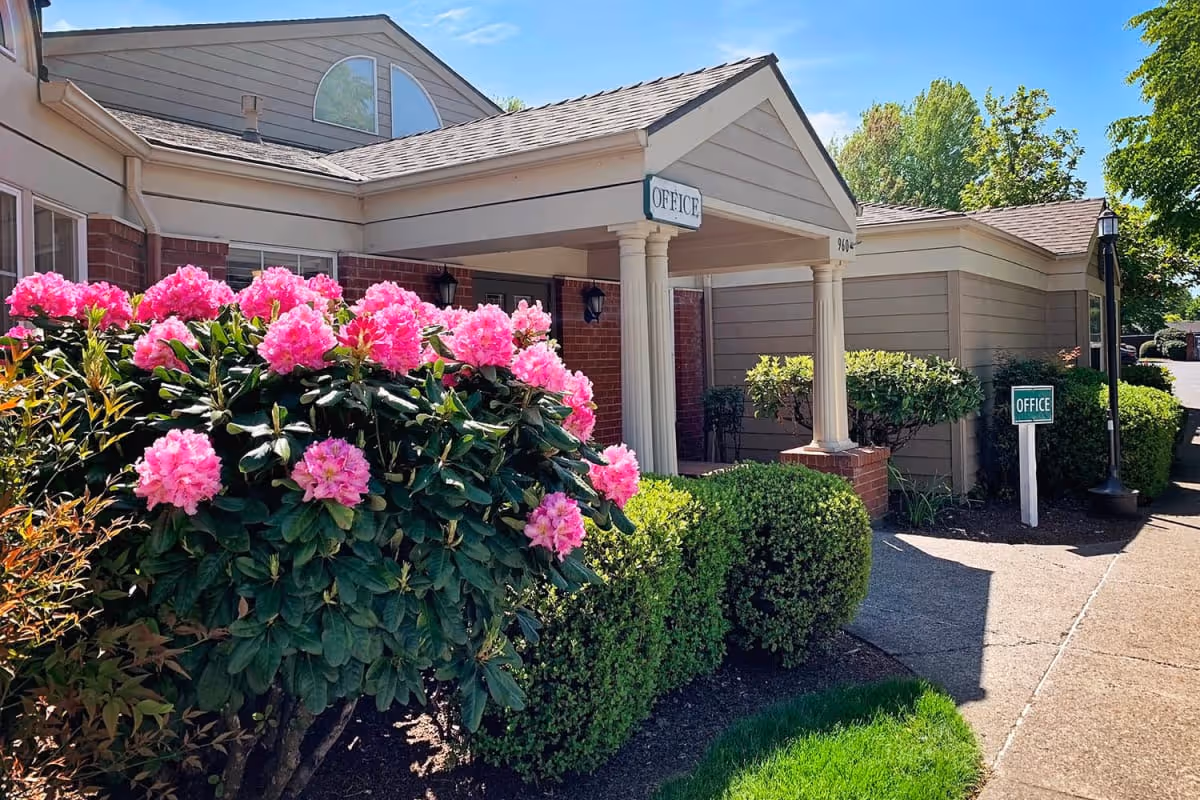 Exterior view of the entrance to a building with a covered porch supported by columns. There are vibrant pink flowers and green bushes in the landscaped area near the walkway. Signs indicating the office are visible near the entrance. The sky is clear and blue.
