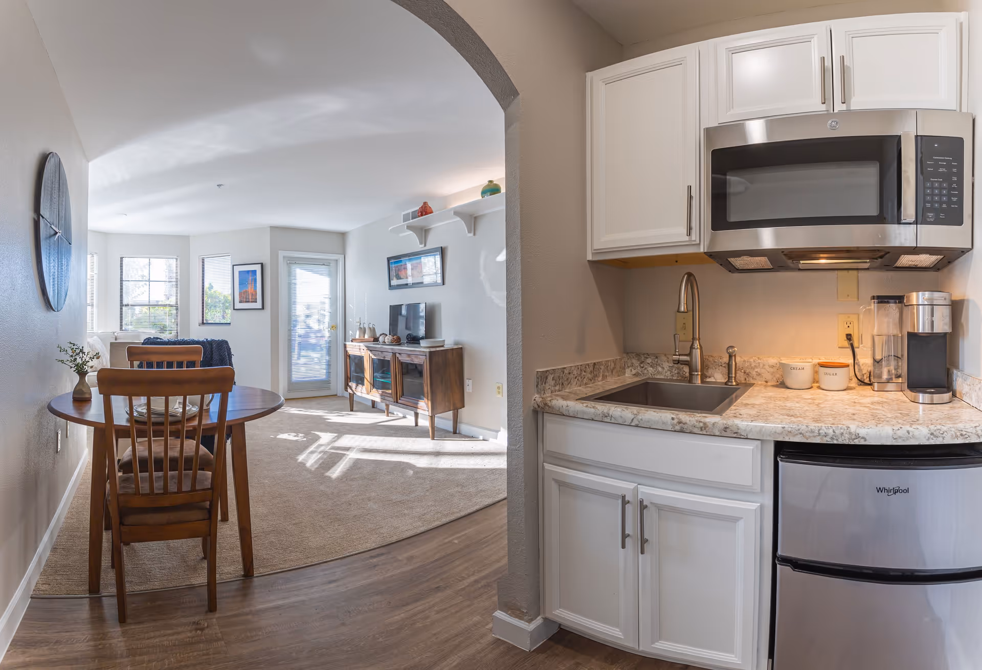 Interior view of a senior living facility showing a small kitchen area with white cabinets, a microwave, a sink, and a mini refrigerator. Adjacent to the kitchen is a dining area with a round wooden table and two chairs. In the background, there is a living room with a TV on a wooden stand, wall art, and a door leading outside. The space is well-lit with natural light from multiple windows.