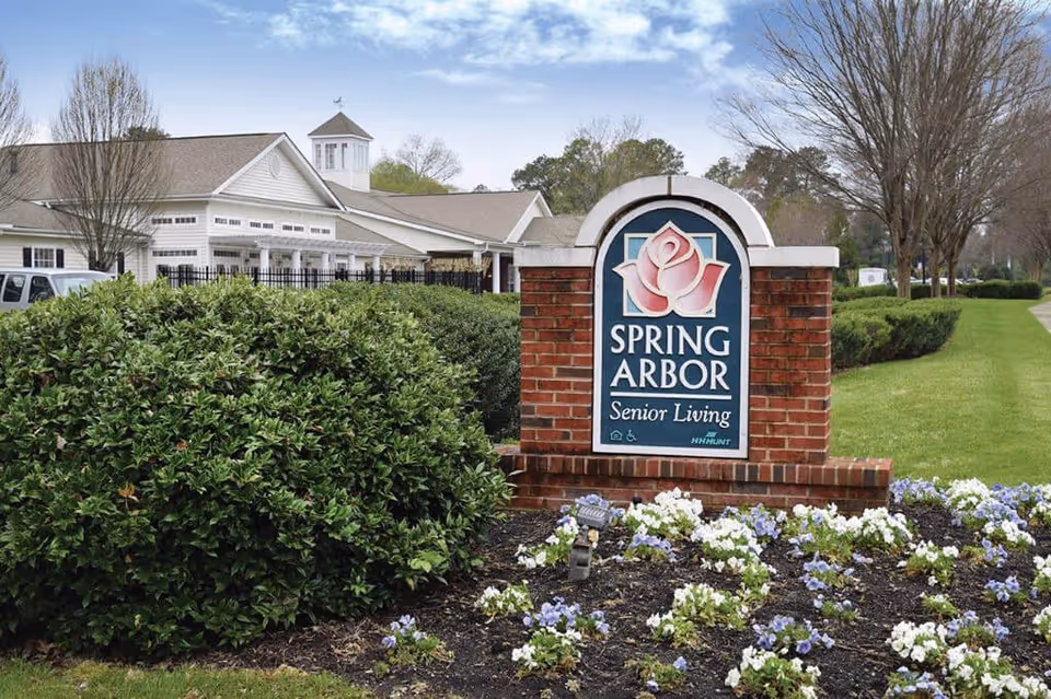 Outdoor view of the entrance sign for Spring Arbor Senior Living, featuring a brick structure with a blue and pink rose logo. The background shows a white building with a cupola, green bushes, a flower bed with white and purple flowers, and a grassy lawn with trees.