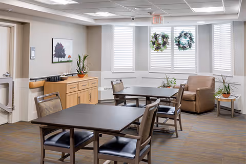 Communal dining room with two tables and chairs, a sideboard, and a seating nook by windows decorated with wreaths.