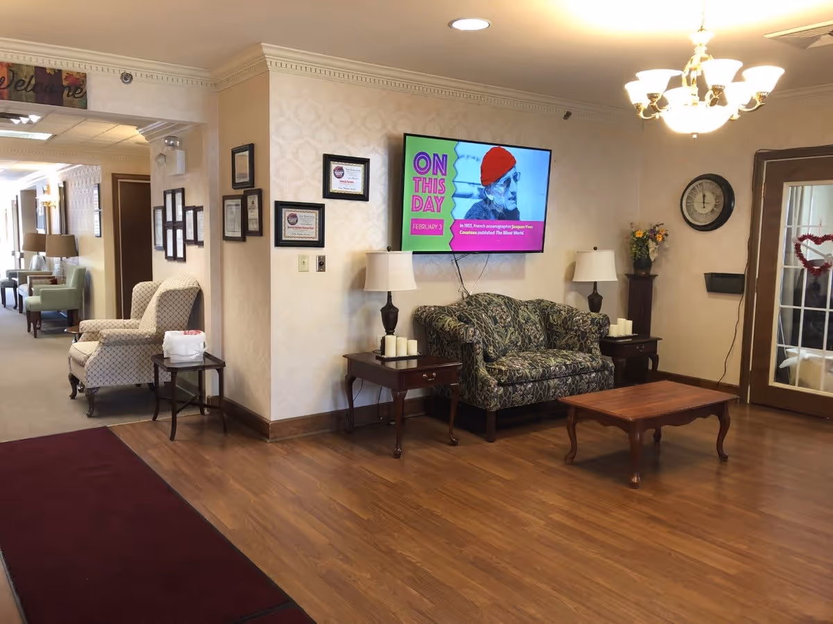 A cozy senior living facility common area with a patterned loveseat, two side tables with lamps and candles, a wooden coffee table, and a wall-mounted TV displaying a colorful 'On This Day' screen. The room has wood flooring, beige wallpaper, framed certificates on the wall, a clock, and a glass door decorated with a heart-shaped wreath. There are additional chairs and lamps visible down a hallway to the left.