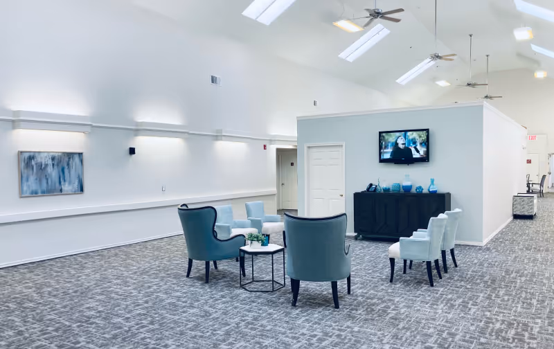 Spacious, skylit assisted-living lounge with a cluster of blue-green armchairs and small tables facing a wall-mounted TV.