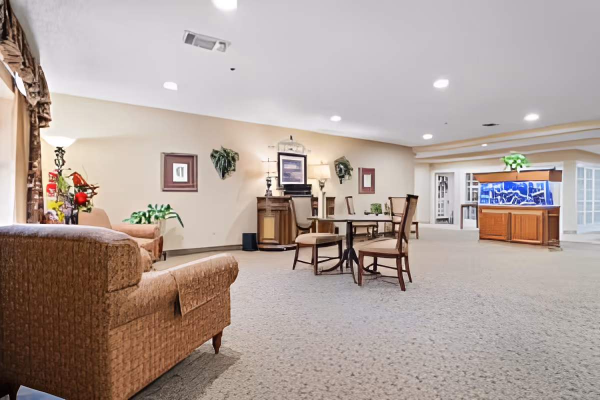 A spacious senior living facility common area with beige carpet and walls, featuring a seating area with upholstered armchairs, a small table with chairs, decorative plants, framed artwork on the walls, and a large fish tank on a wooden stand.