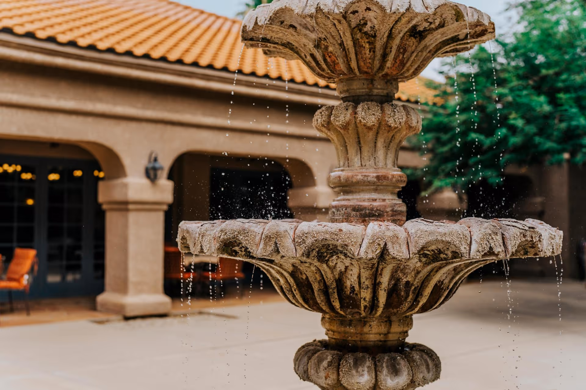 Close-up view of a two-tiered stone water fountain with water gently flowing from the top tier to the bottom tier, set in an outdoor courtyard area with a building featuring arches and a tiled roof in the background.