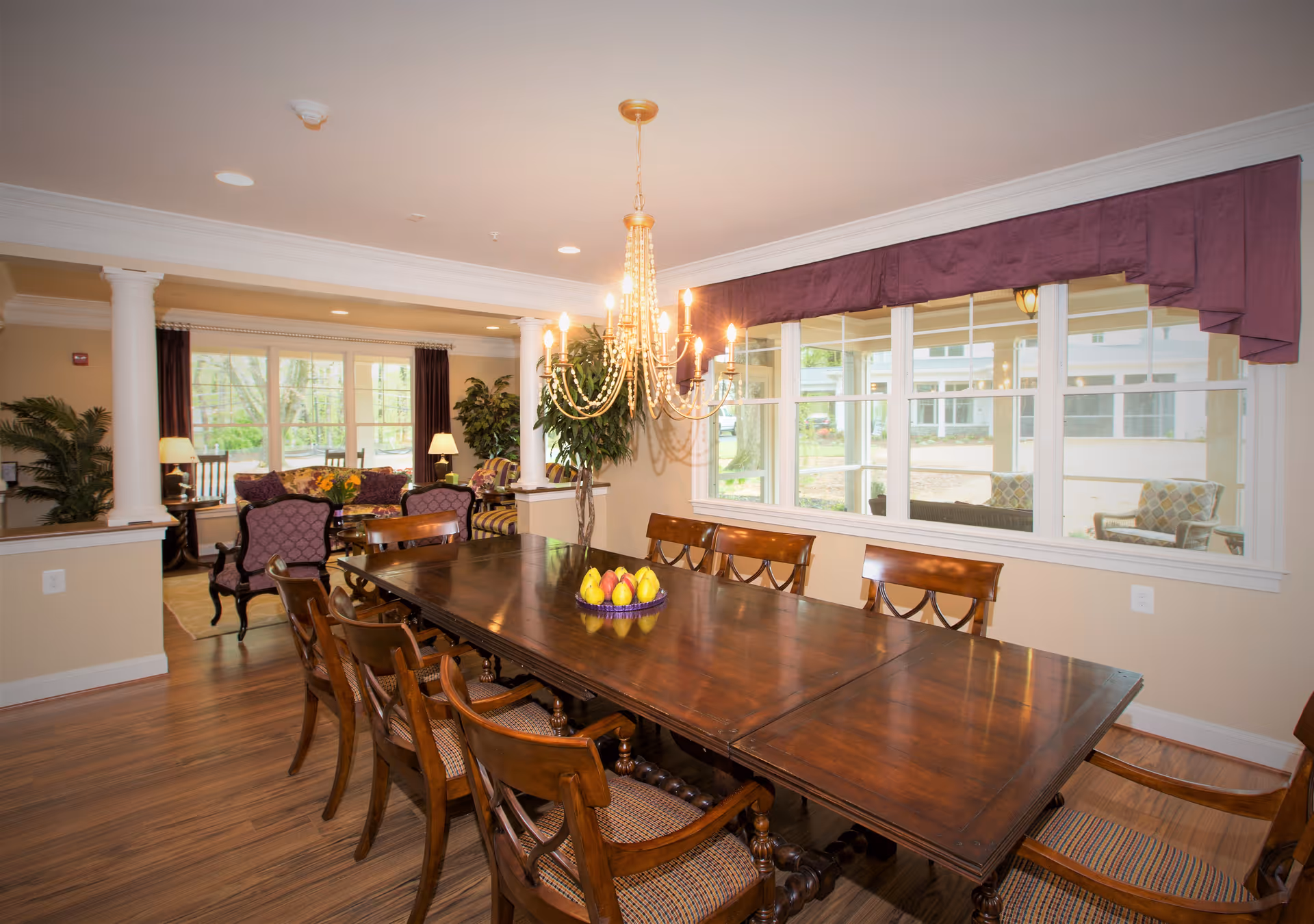 A spacious dining room with a large wooden dining table surrounded by wooden chairs with cushioned seats. A chandelier with multiple lights hangs above the table. The room has large windows with a purple valance, allowing natural light to brighten the space. In the background, there is a cozy living area with armchairs, a sofa, lamps, and indoor plants. The flooring is wooden, and the walls are painted in a warm beige tone.