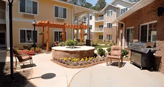 Outdoor courtyard area of a senior living facility with a circular brick fire pit surrounded by flower beds, patio chairs, a barbecue grill, and a wooden pergola. The courtyard is enclosed by multi-story residential buildings with windows and balconies.