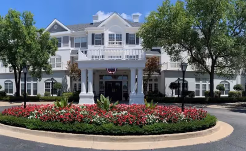 Front exterior view of a large white senior living facility building with a covered entrance supported by columns, surrounded by trees and a circular flower bed with red and white flowers in front.