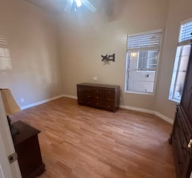 Empty room with light wood flooring, beige walls, a ceiling fan with light, two windows with blinds, and three pieces of dark wood furniture including a dresser, a nightstand with a lamp, and a tall cabinet.