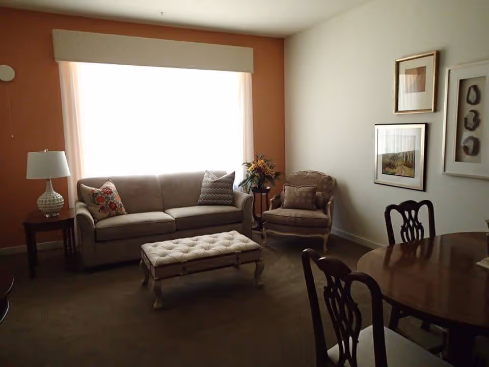 A cozy living room with a beige sofa adorned with two decorative pillows, a matching armchair with a cushion, and a tufted ottoman in front of the sofa. There is a wooden side table with a white lamp on the left side of the sofa, a round wooden dining table with chairs on the right, and framed artwork hanging on the white wall. The room has a large window with sheer curtains letting in natural light, and a small table with a flower arrangement beside the armchair.