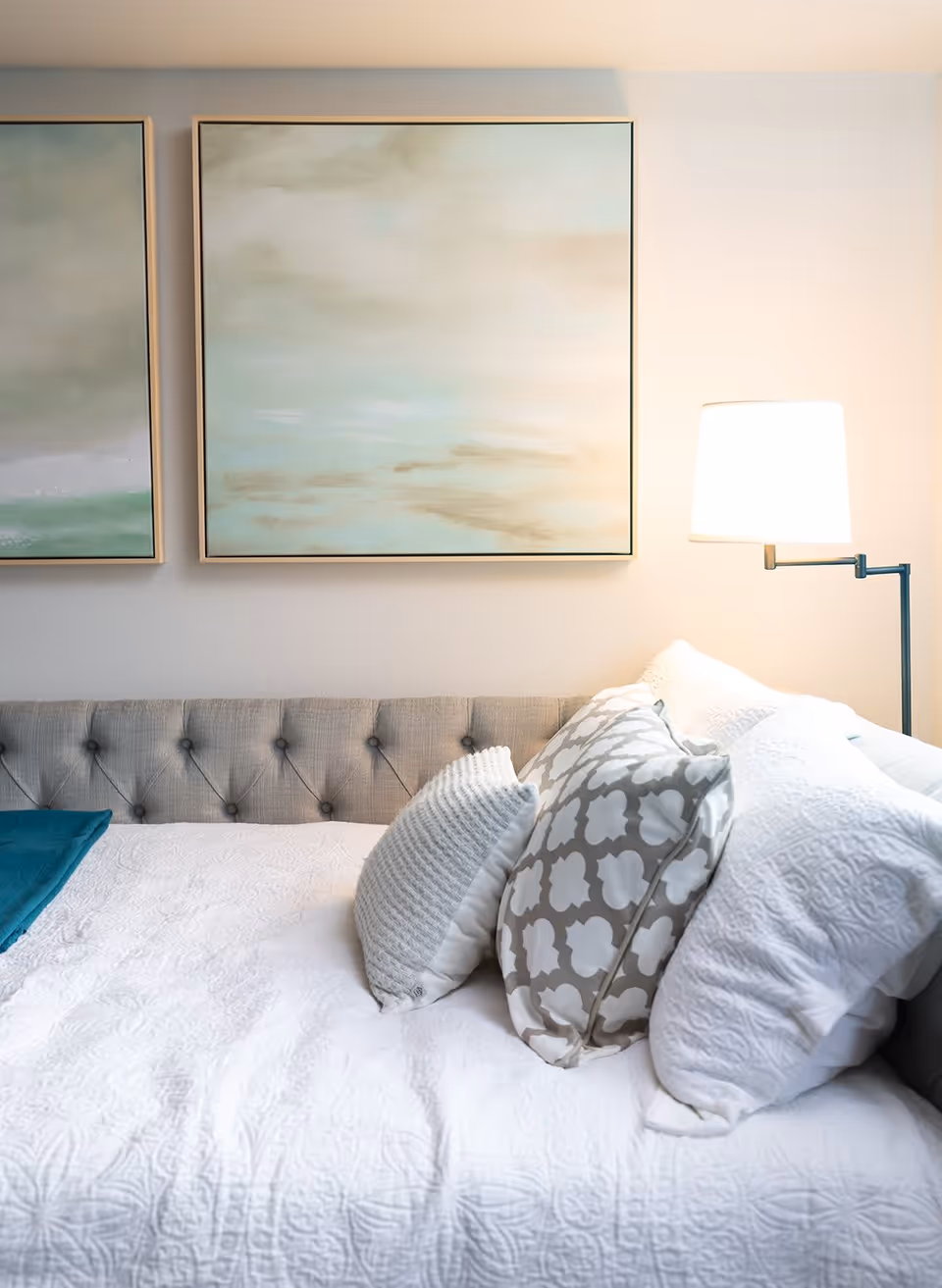 A cozy bed with a tufted gray headboard, white textured bedding, and several decorative pillows in white and gray patterns. A modern floor lamp with a white shade is positioned next to the bed, and two abstract landscape paintings with soft green and beige tones hang on the wall above the headboard.
