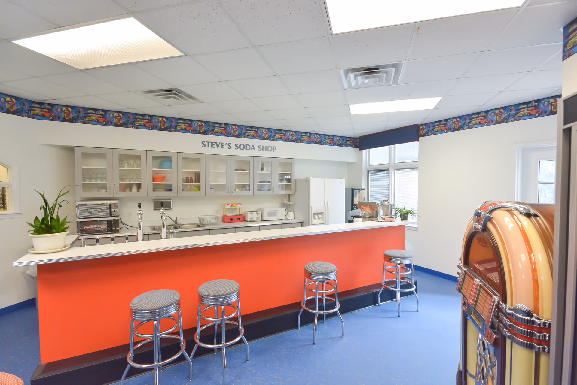 Interior view of a retro-style soda shop area named 'Steve's Soda Shop' with a long counter painted red, five metal stools with cushioned seats, cabinets with glass doors displaying dishes and cups, a refrigerator, and a vintage jukebox on the right side. The room has a blue floor and a ceiling with fluorescent lights.