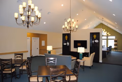 Interior view of a spacious common area in a senior living facility with multiple seating arrangements including tables with chairs and armchairs. The room features high vaulted ceilings with multiple chandeliers, a TV mounted on a black shelving unit, and large windows allowing natural light in the background.