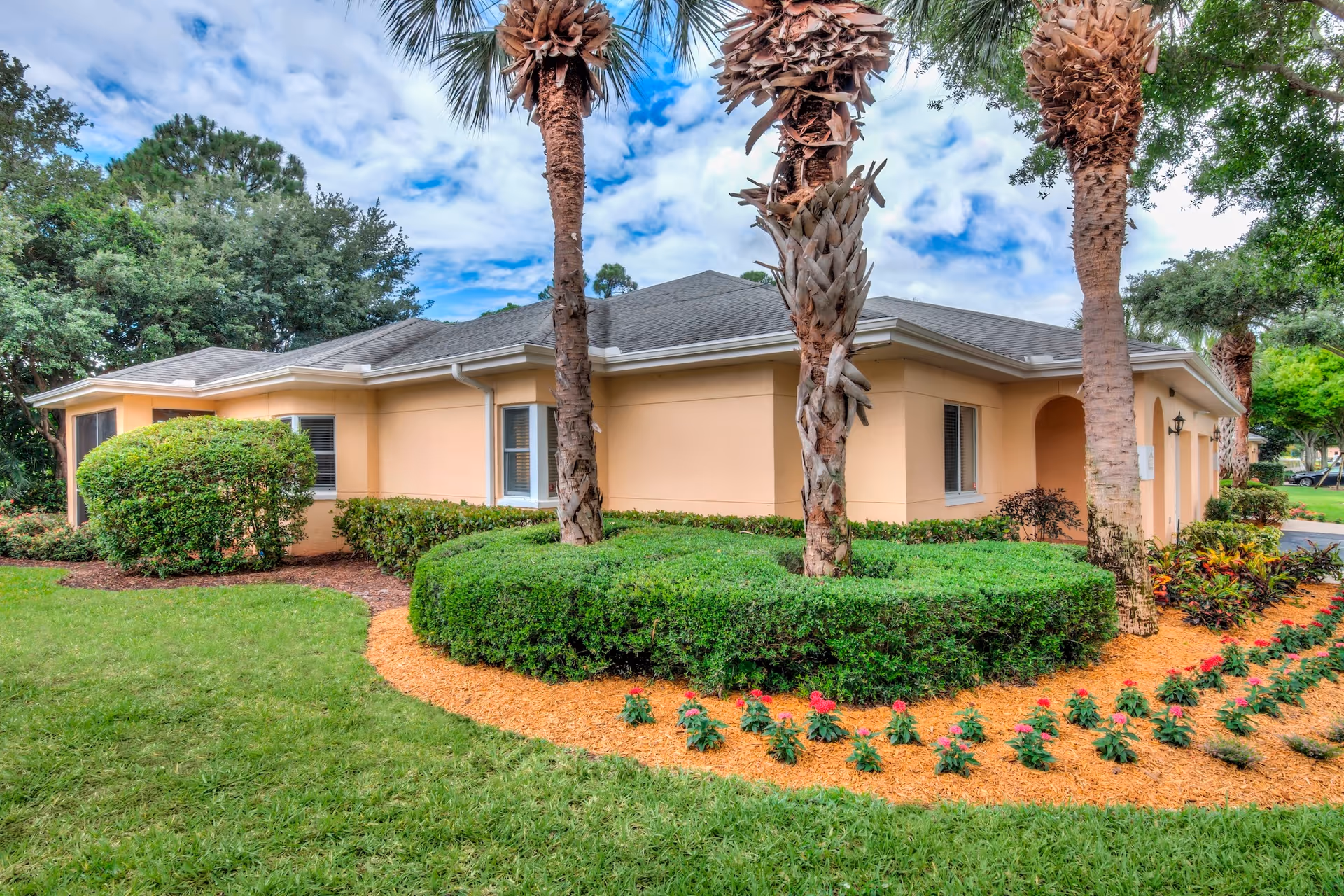 Exterior view of a single-story building with beige walls and a gray roof, surrounded by neatly trimmed bushes, palm trees, and a flower bed with red flowers. The sky is partly cloudy.