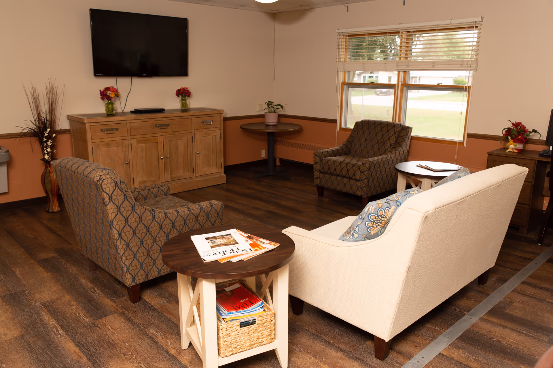 A cozy living room area with two patterned armchairs and a beige sofa with a decorative pillow. There is a round wooden side table with magazines and a basket underneath, a wooden cabinet with two flower vases and a flat-screen TV mounted on the wall above it. A window with blinds lets in natural light, and there is a small round table with a potted plant in the corner. The room has wooden flooring and warm-toned walls.
