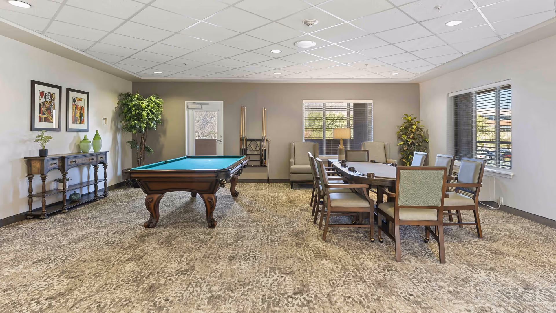 A spacious recreational room with a pool table in the center, a long wooden table with eight chairs on the right, two framed artworks on the left wall above a wooden console table with decorative vases, two large windows with blinds, a door, and two potted plants in the corners.