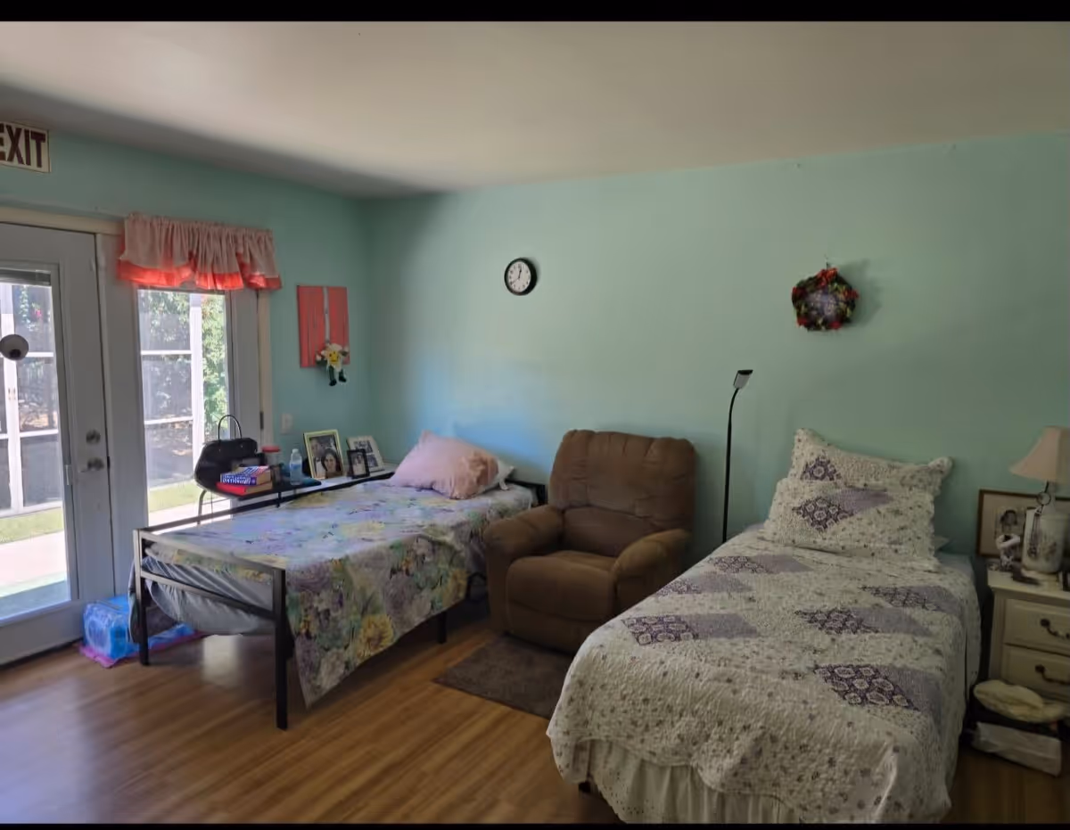 A cozy room with two single beds, one with floral bedding and the other with a white and purple quilt. Between the beds is a brown recliner chair and a floor lamp. There is a small nightstand with a lamp and framed photos on the right side. A clock and a decorative wreath hang on the light blue wall. A glass door with an exit sign above it lets in natural light and shows greenery outside.
