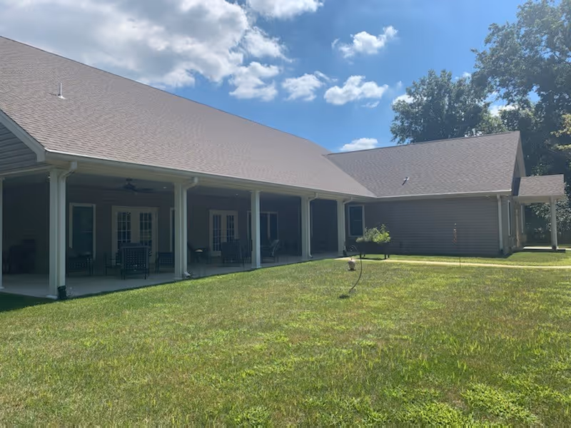 Exterior view of a single-story building with a covered porch supported by white columns. The porch has several chairs and tables. The building is surrounded by a grassy lawn under a partly cloudy sky.