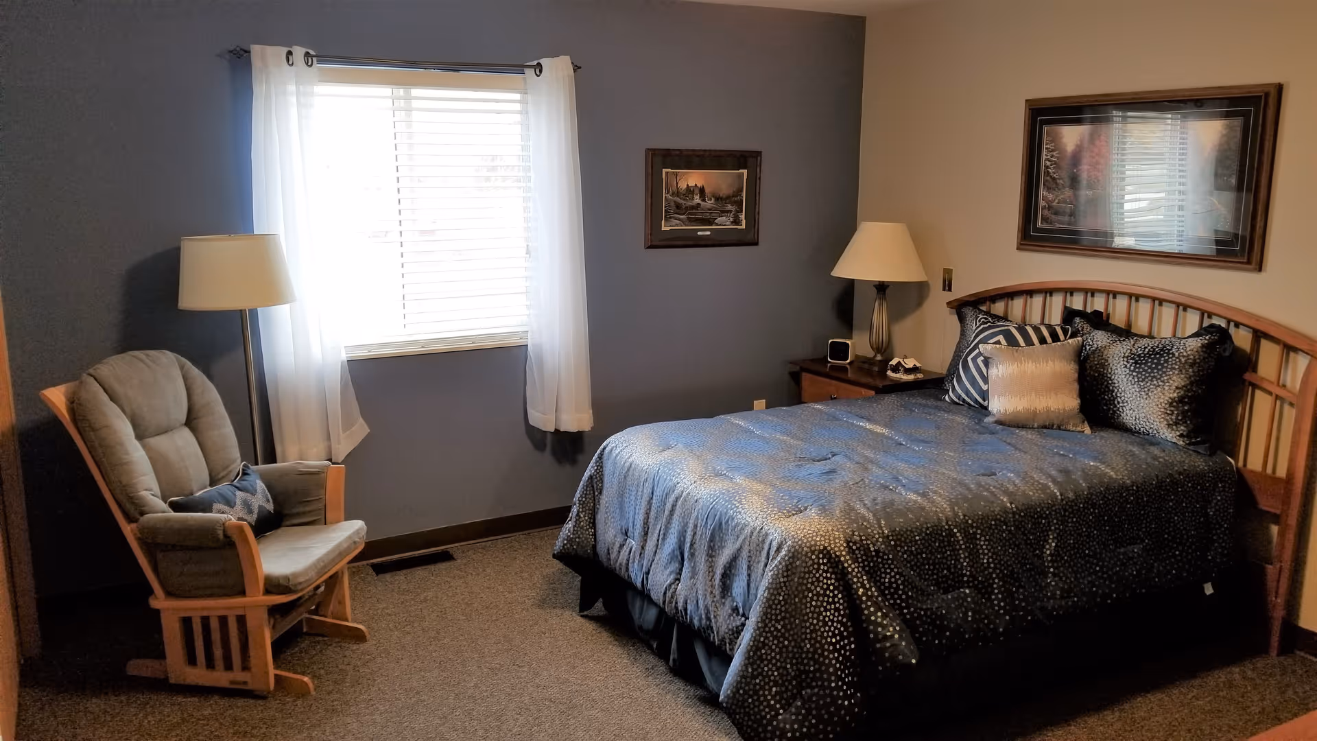 A cozy bedroom with a double bed covered in a dark blue comforter with decorative pillows. There is a wooden rocking chair with cushions and a small pillow next to a floor lamp by a window with white curtains. Two framed pictures hang on the walls, and a wooden nightstand with a lamp and a clock is beside the bed.