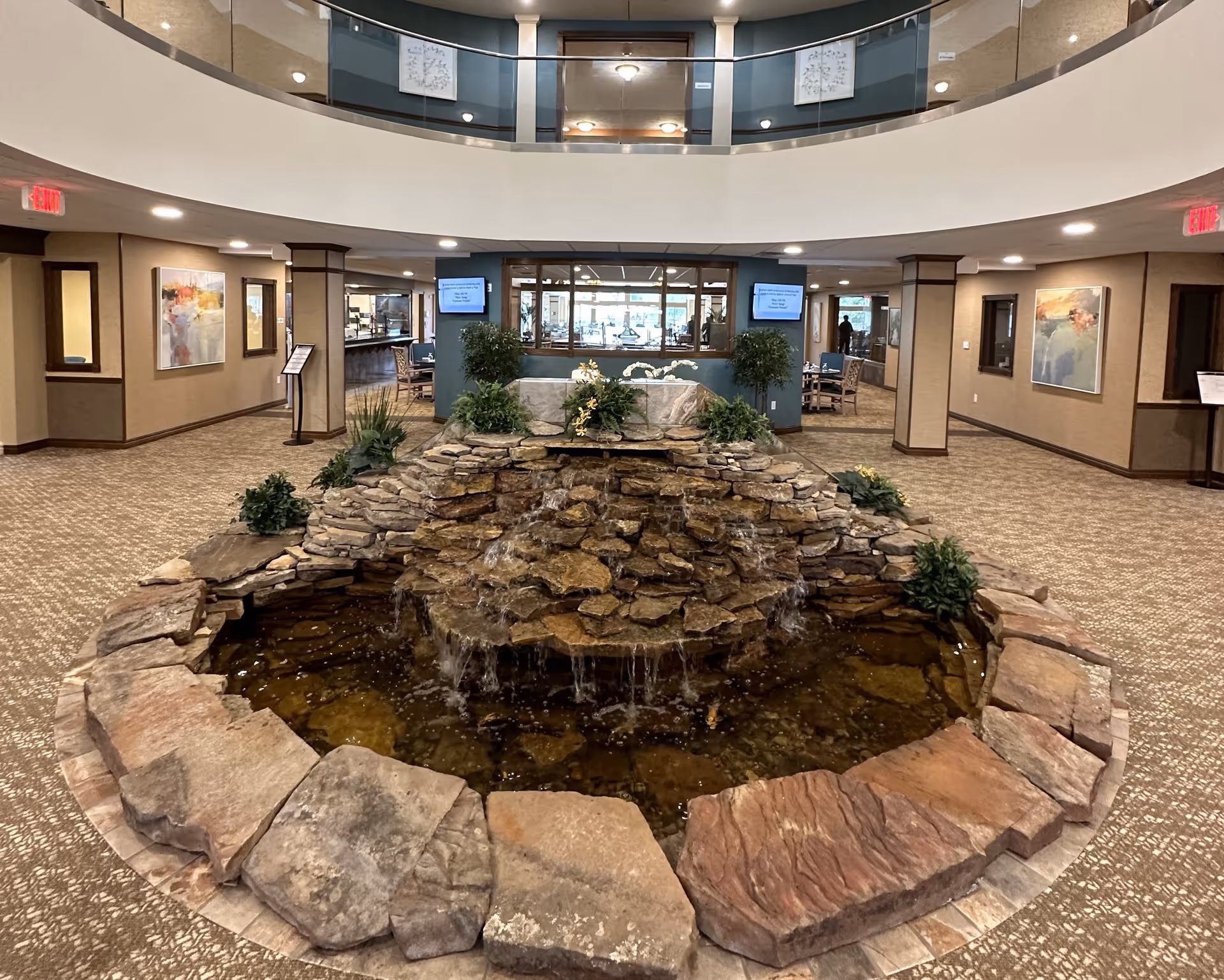 Indoor area of a senior living facility featuring a central stone water fountain with cascading water surrounded by plants. The space has carpeted floors, beige walls with artwork, and a second-floor balcony with glass railing overlooking the area. There are seating areas and digital screens visible in the background.