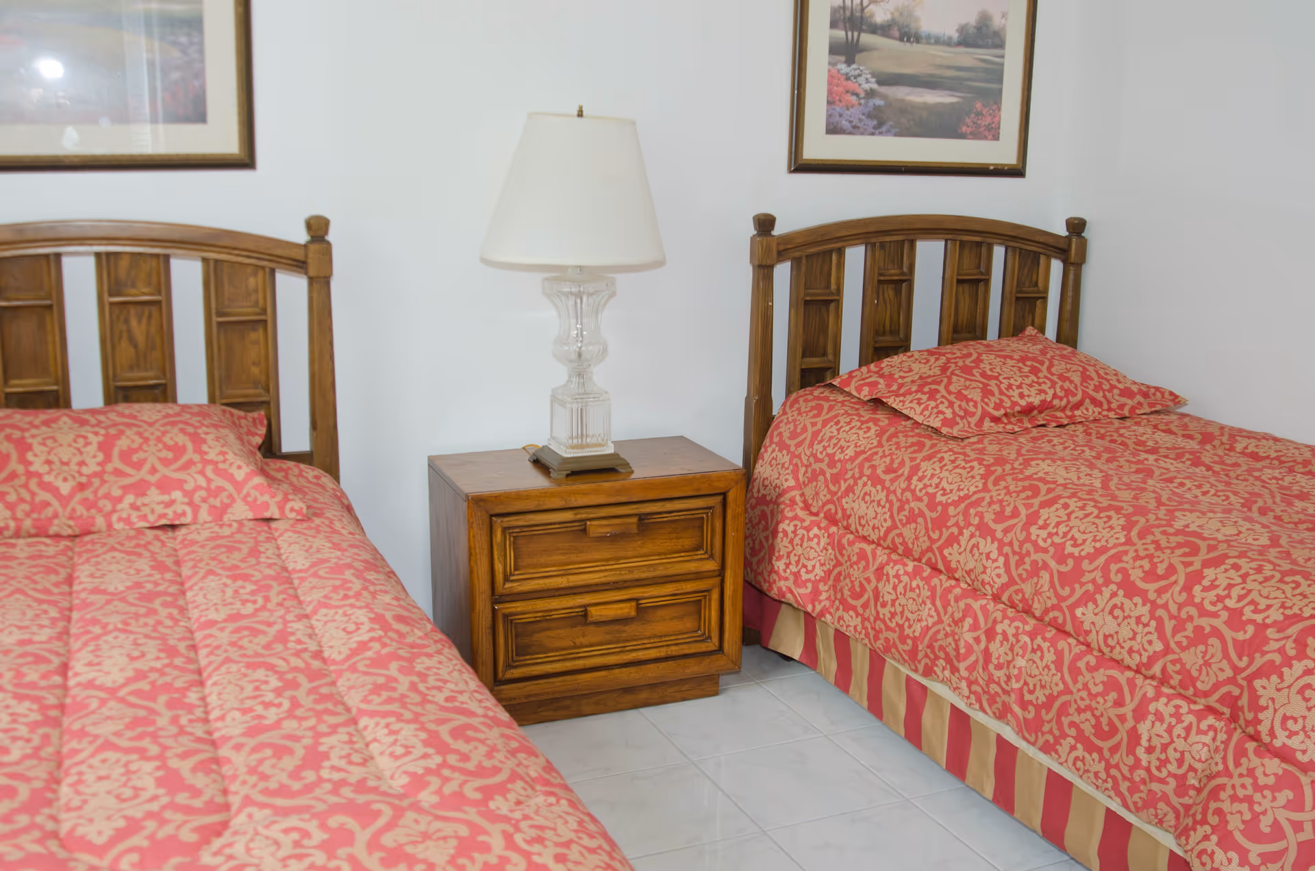 A bedroom with two single beds featuring wooden headboards and red patterned bedspreads. Between the beds is a wooden nightstand with two drawers and a clear glass lamp with a white lampshade. Two framed landscape paintings hang on the white walls above the beds.