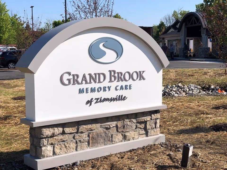 Outdoor stone and white sign for Grand Brook Memory Care of Zionsville with a logo above the text, situated on a landscaped area with some trees and a building in the background.