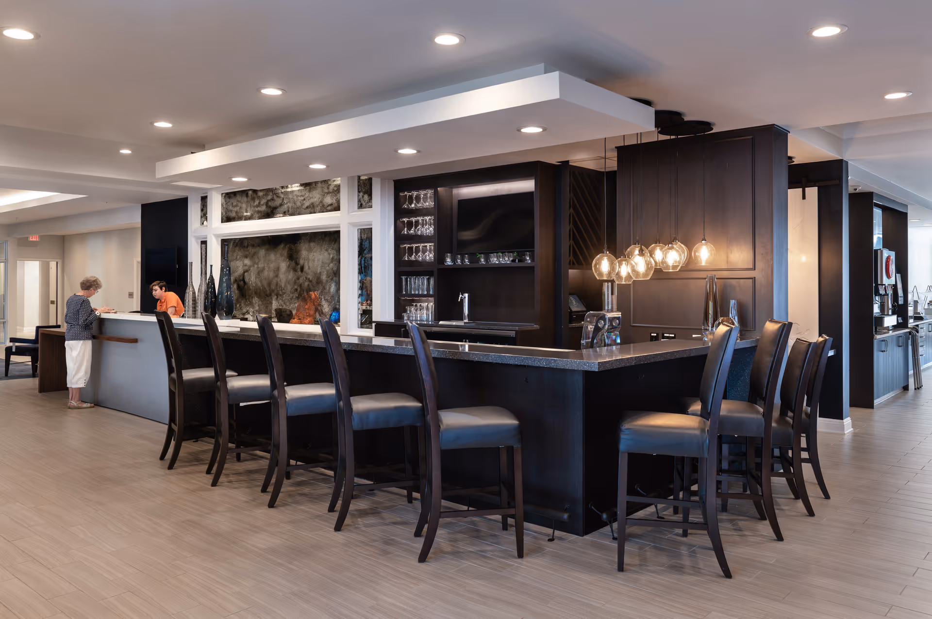 Contemporary communal bar and lounge area with a long dark counter, multiple bar stools, pendant lights, and two people at the service desk.