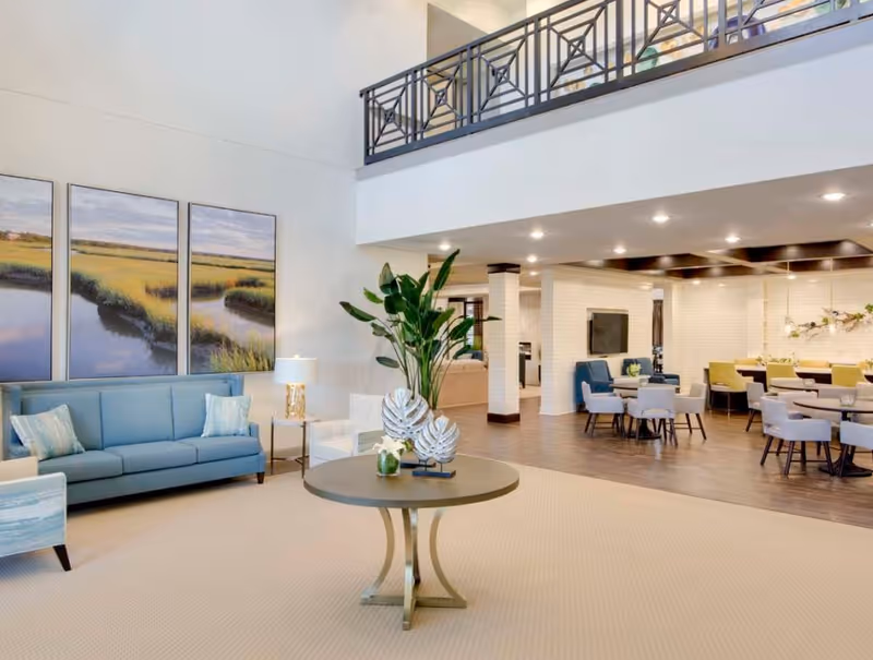 A spacious and well-lit common area in an assisted living facility featuring a blue sofa, armchairs, a round table with decorative plants, and a dining area with multiple tables and chairs. The room has high ceilings, large landscape paintings on the wall, and a mezzanine with a decorative railing.
