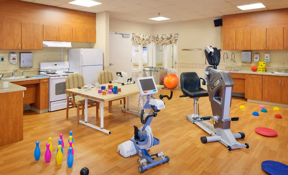 Well-lit interior activity/therapy room with exercise bikes, a kitchenette, an activity table, and colorful therapy toys on a wood floor.
