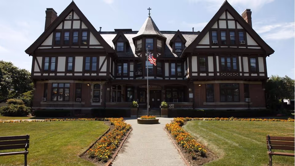 A Tudor-style senior living facility with a central entrance and manicured flower beds along a paved walkway.