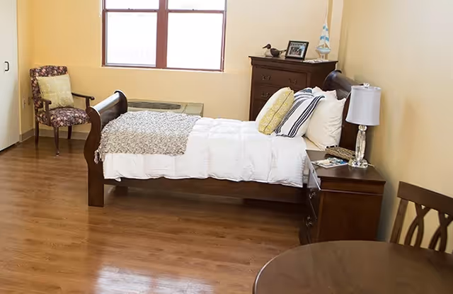 Simple bedroom with a wooden bed dressed in white linens, a nightstand with lamp, dresser, and chairs on wood flooring.