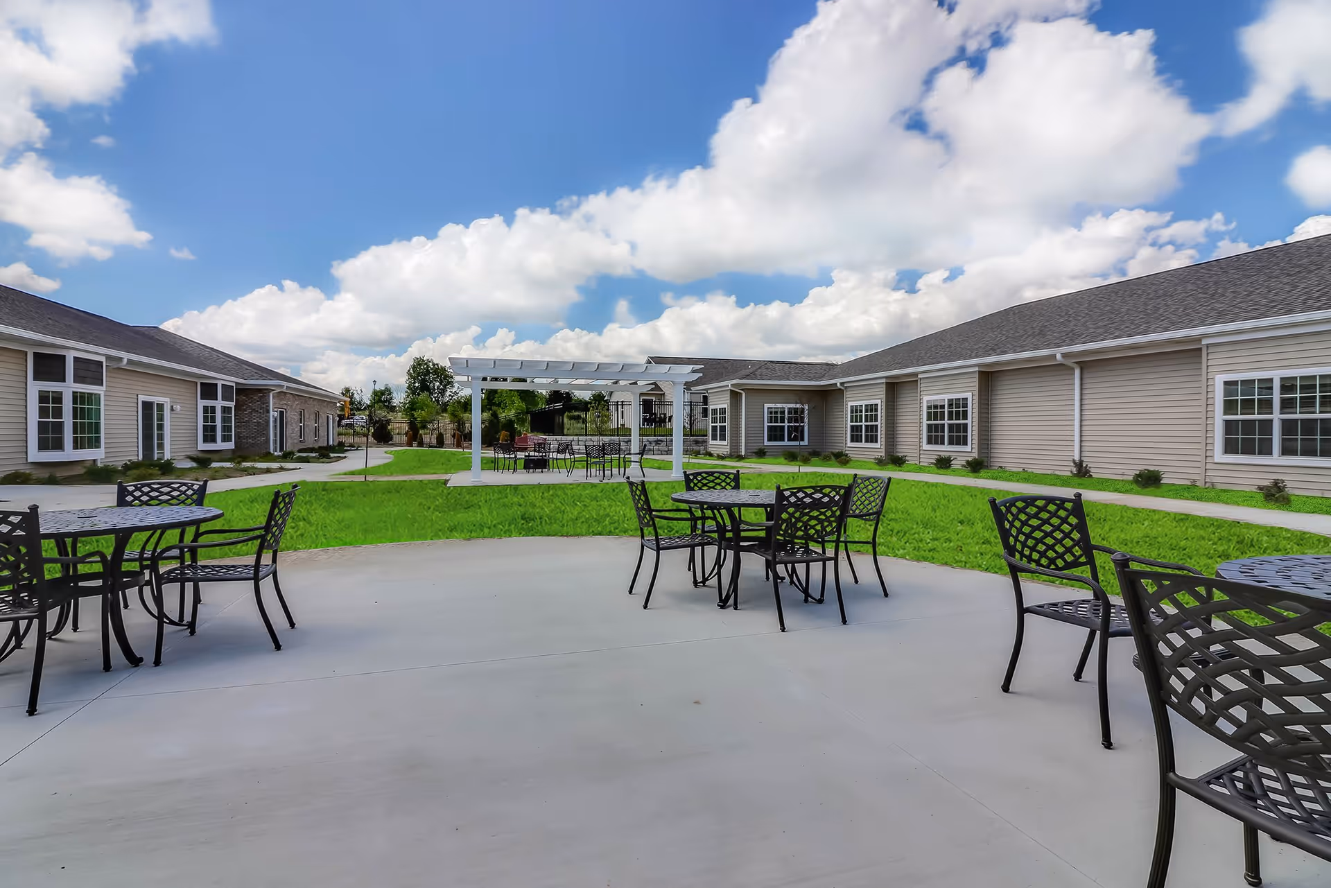 Outdoor patio area at Springvale Assisted Living & Memory Care with several black metal tables and chairs on a concrete surface, surrounded by green grass and beige single-story buildings under a partly cloudy blue sky.
