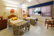 A bright shared patient room with two hospital beds, bedside tables, a curtain divider, chairs and a large window with a wall-mounted TV.