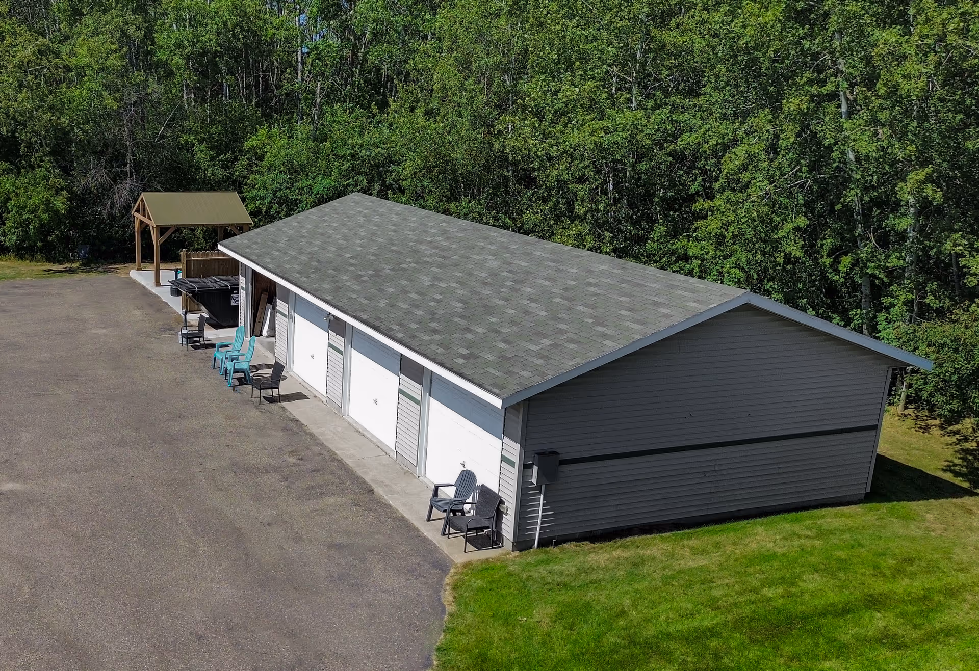 A long single-story garage building with multiple white roll-up doors, chairs along the front, a paved driveway and trees in the background.