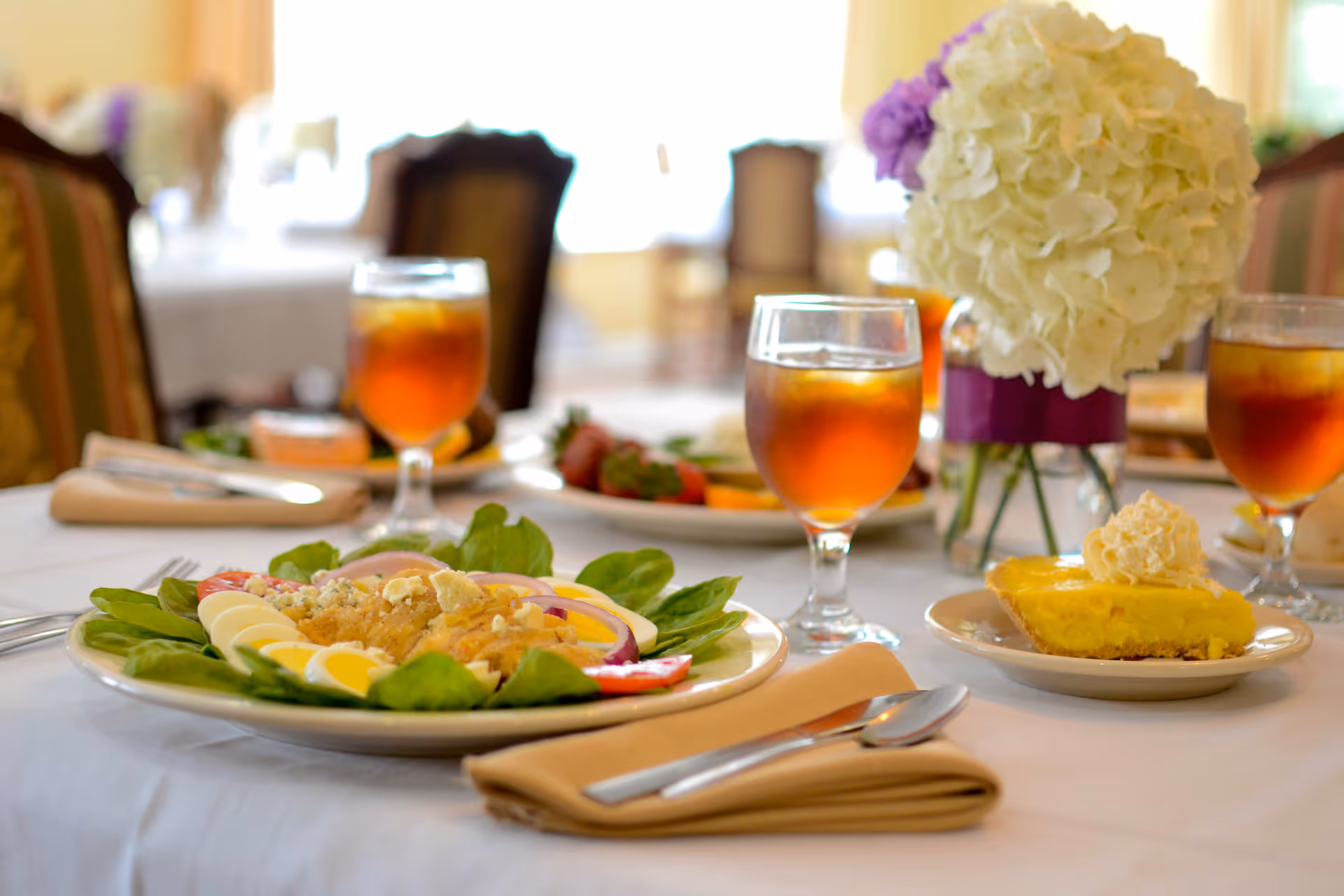 A dining table set with plates of salad and dessert, glasses of iced tea, beige napkins with silverware, and a centerpiece of white and purple flowers in a vase, in a softly lit dining room.