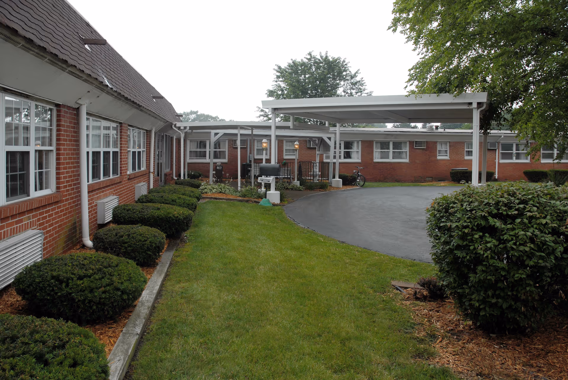 Brick single-story senior care facility exterior with a covered circular driveway, manicured shrubs, and windows.