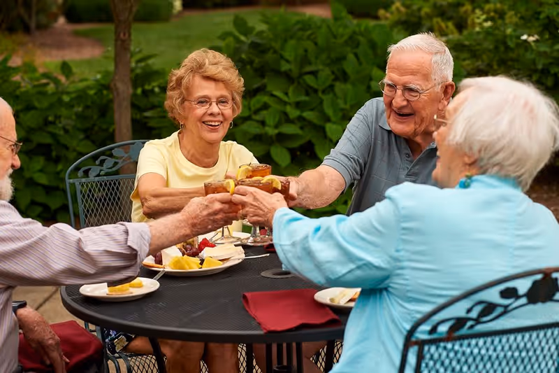 Four elderly people sitting around a black metal outdoor table, smiling and clinking glasses of iced tea with lemon slices. The table has plates with food and red napkins. They are in a garden setting with green bushes and trees in the background.