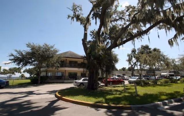 Exterior view of a two-story building with a parking lot in front, several cars parked, and large trees with hanging moss surrounding the area under a clear blue sky.