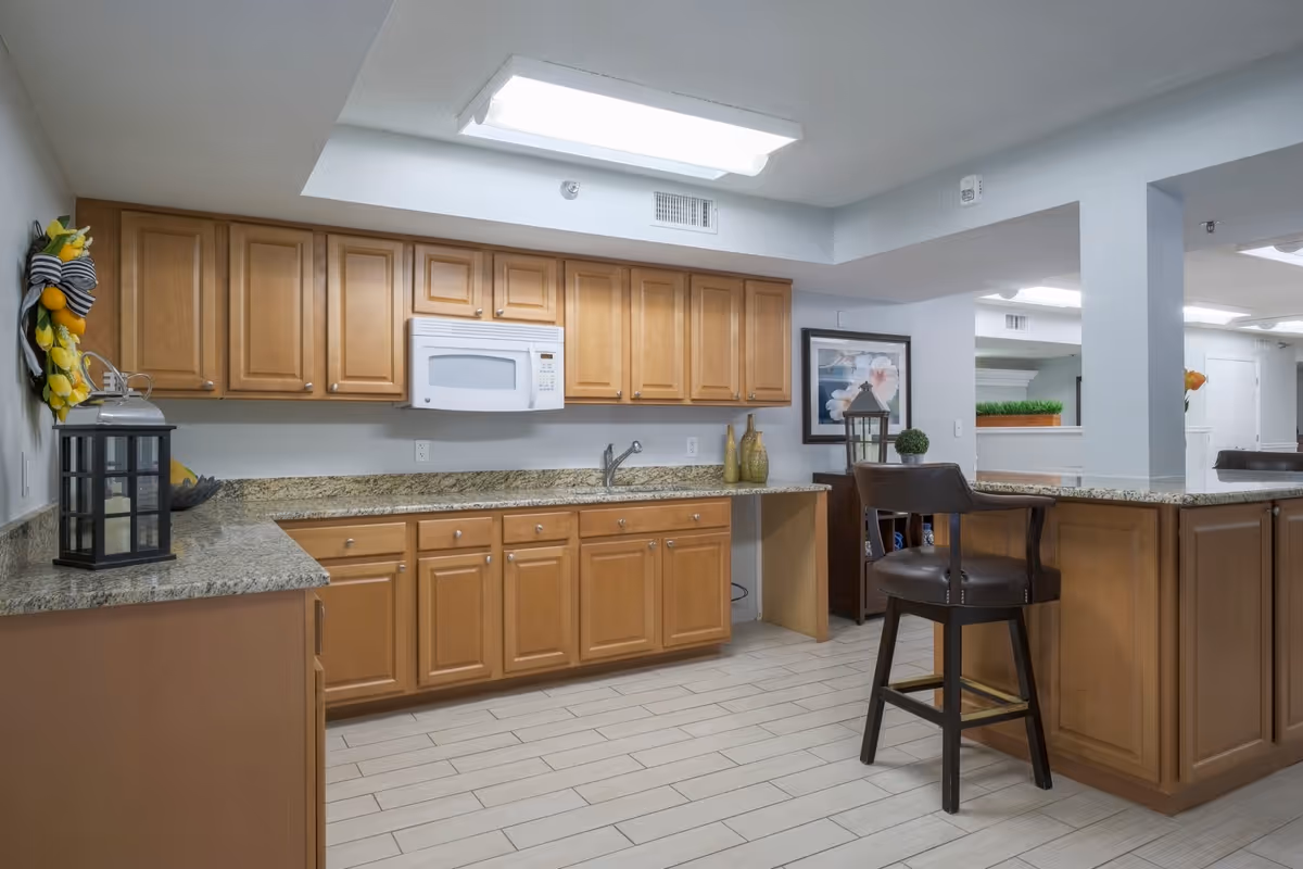 Interior view of a kitchen area in a senior living facility with wooden cabinets, granite countertops, a white microwave, a sink, and a high chair. The space is well-lit with ceiling lights and decorated with a wreath, vases, and framed artwork.