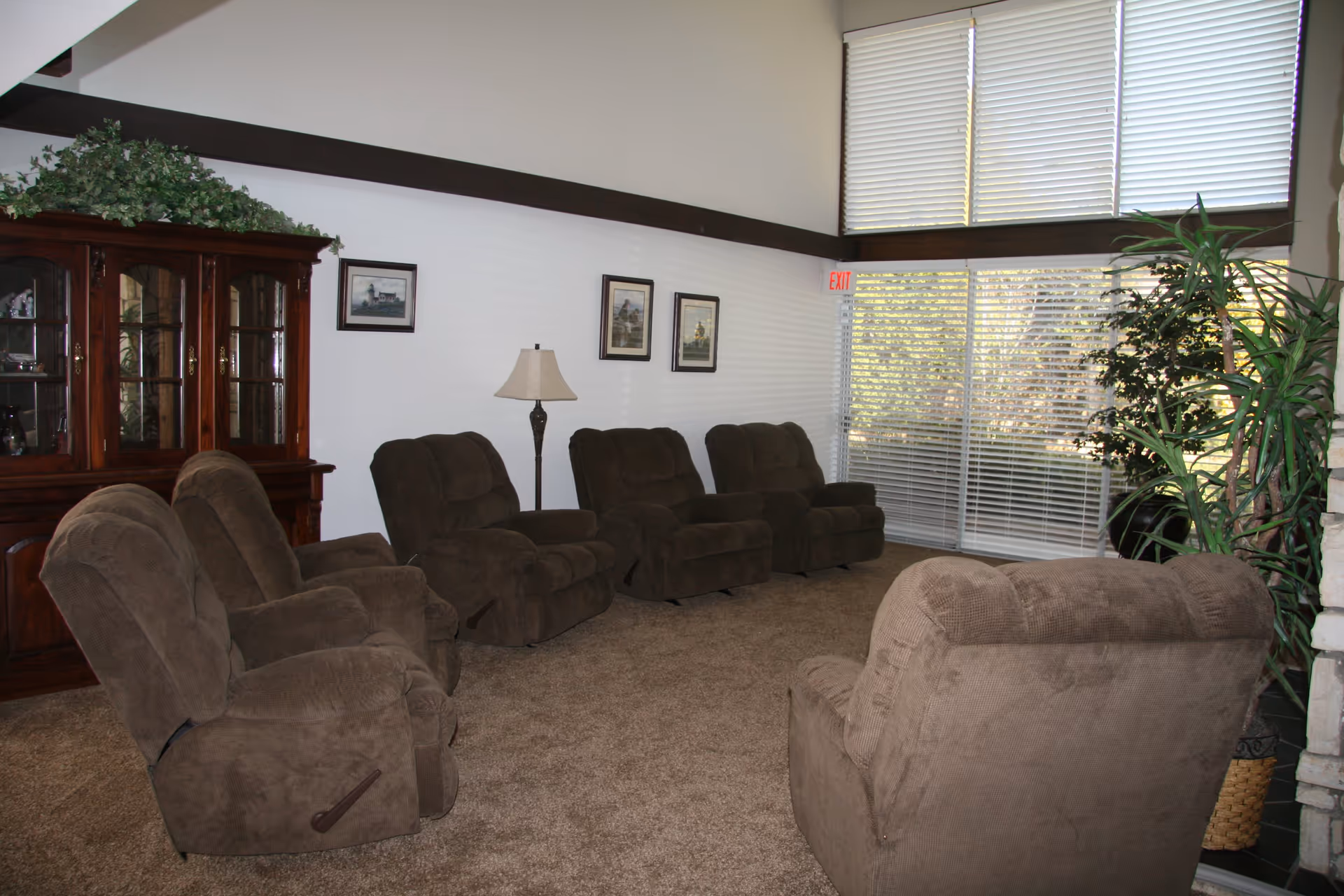 A cozy living room with six brown recliner chairs arranged in a semi-circle on a carpeted floor. The room features a wooden cabinet with glass doors on the left, a floor lamp, three framed pictures on the wall, large windows with white blinds, and several green plants near the windows.