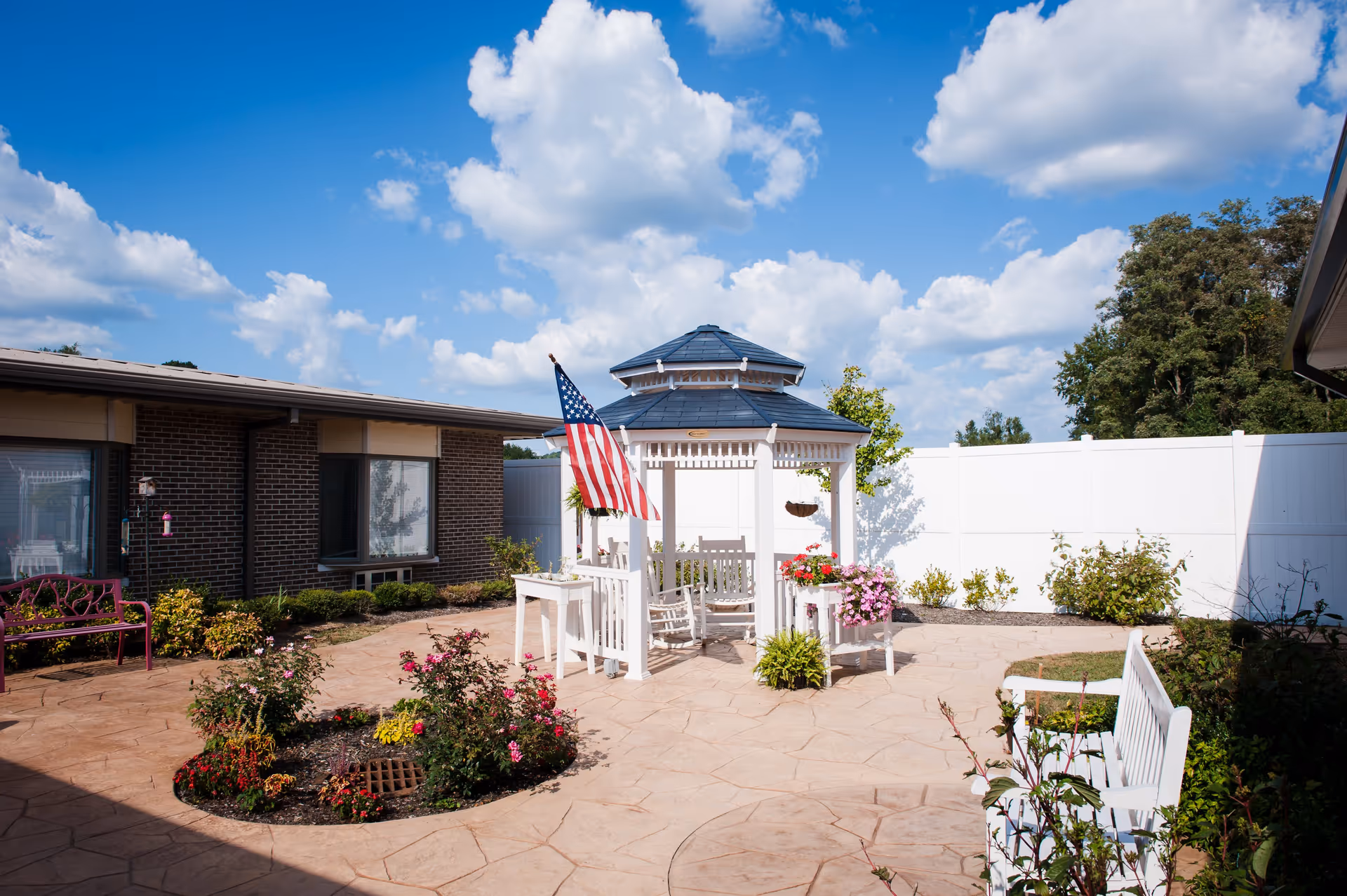 Outdoor courtyard area with a white gazebo featuring a blue roof and an American flag. The courtyard has a paved stone floor, flower beds with colorful flowers, white benches, and a brick building in the background under a partly cloudy blue sky.