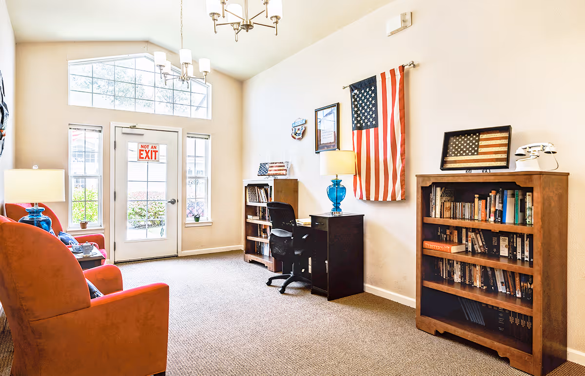 A bright room with beige walls and carpet featuring two orange armchairs with blue lamps on side tables, a desk with a black office chair, two wooden bookshelves filled with books, and American flags displayed on the wall and on one bookshelf. A door with a 'Not an Exit' sign and windows letting in natural light are visible.