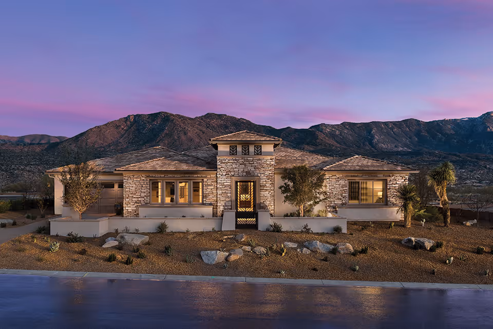 Exterior front view of a single-story stone and stucco building with a tiled roof, surrounded by desert landscaping with rocks, small plants, and trees, set against a backdrop of mountains under a purple and pink sunset sky.