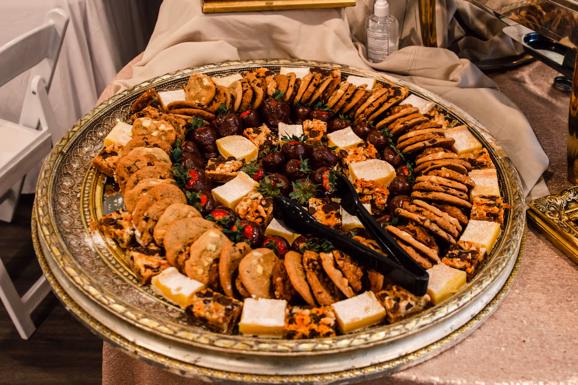A large ornate silver tray filled with an assortment of cookies, chocolate-covered strawberries, and various small dessert bars arranged in concentric circles, with black serving tongs placed on top. The tray is set on a beige tablecloth with a white folding chair visible in the background.