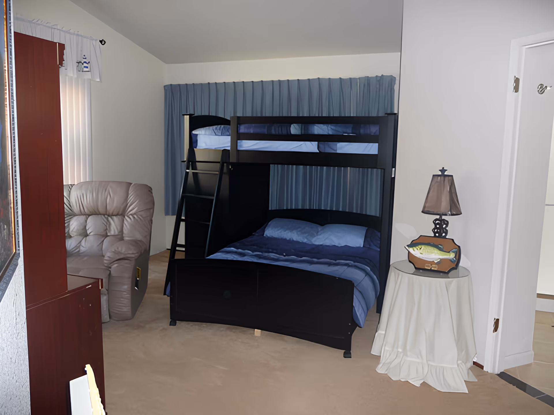 A bedroom with a black bunk bed featuring a double bed on the bottom and a single bed on top, both made with blue bedding. To the left of the bunk bed is a beige recliner chair. A small round table covered with a white cloth holds a lamp and a decorative fish. The room has beige carpet and light-colored walls with a window covered by blue curtains behind the bunk bed.