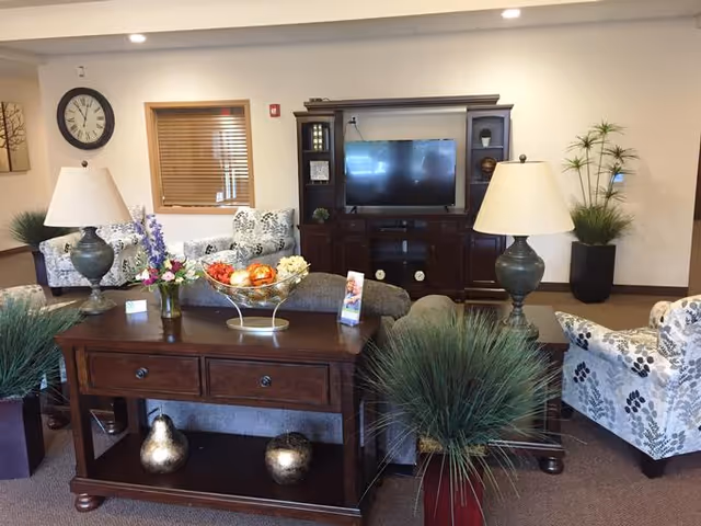 A cozy living room area with a dark wooden TV cabinet holding a flat-screen television, flanked by two table lamps with beige shades. There are two floral-patterned armchairs and a sofa with a floral design. A wooden console table in the foreground holds decorative items including a bowl of artificial fruit and a vase of flowers. Several potted plants are placed around the room, and a round wall clock is visible on the left wall above a window with wooden blinds.