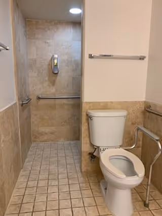 Accessible bathroom with beige tiled floor and walls, featuring a white toilet with grab bars on both sides and a walk-in shower area with a wall-mounted soap dispenser and a grab bar.